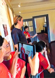 Zwei Schüler schauen auf ein Pad. Auf dem Pad ist ein Mitschüler zu sehen, der etwas vorträgt. Im Hintergrund sind drei Personen, die in Richtung des Vortragenden schauen.