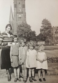 Auf einer schwarz-weiß Fotografie sind in einer Reihe stehend eine Frau mit vier Kindern zu sehen. Im Hintergrund befindet sich ein Kirchturm.