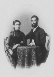An einem Tisch sitzen eine Frau und ein Mann, der seinen Arm um sie legt. Sie trägt ein schlichtes Kleid, er einen Anzug.