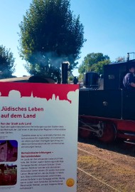 Eine alte Dampflock fährt in Niederzissen ein. Im Vordergrund ist ein Modul der Wander-Ausstellung zu sehen.