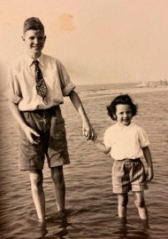 Ein junger Mann in Hemd, Krawatte und kurzer Hose steht am Strand im Wasser mit seinem kleineren Bruder an der Hand.